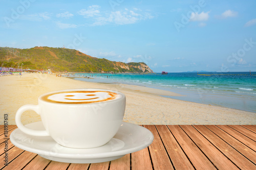 White cup of smile coffee on wooden table with defocused blue sea, white sand beach background.