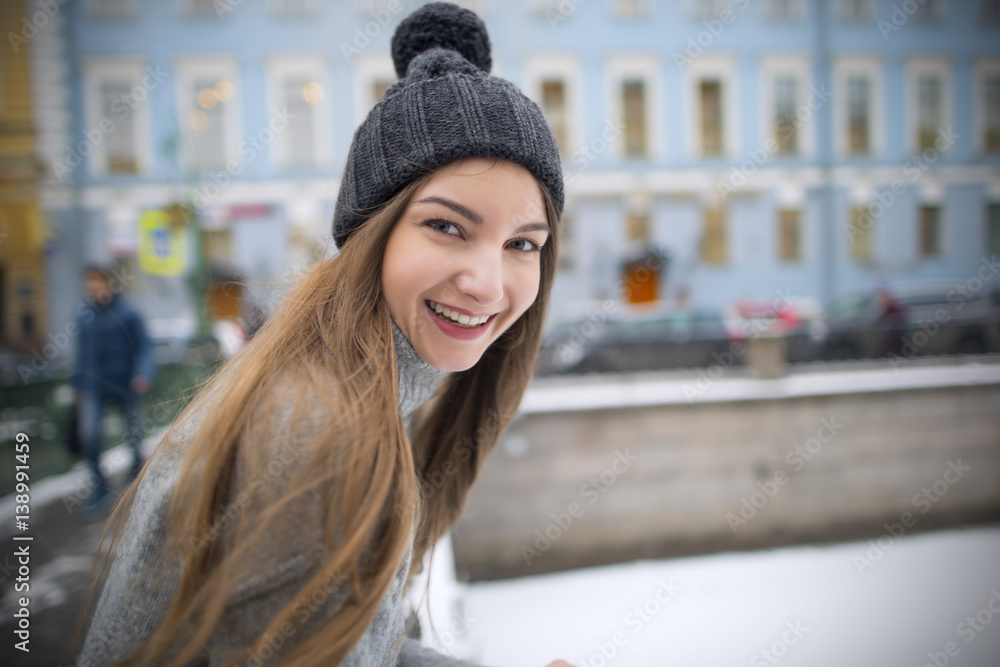 Beautiful girl with long hair in a sweater and a warm hat smiling and posing against the backdrop of views of the city.