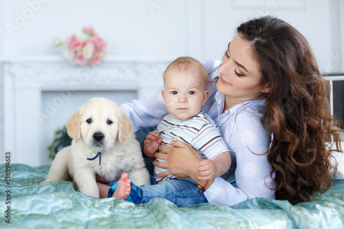 Little boy,barefoot,sitting on the bed  with her mother, a beautiful woman with long curly hair, black hair, wearing a white shirt and blue jeans, along with them - a pet, puppy golden retriever