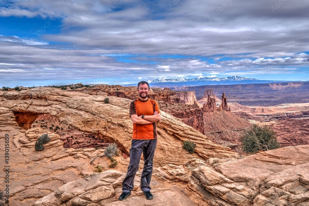 Bearded hipster man smiling on cliff with canyon views. Mesa Arch in Canyonlands National Park. Moab. Cedar City. La Sal Mountains.  Utah. United States.