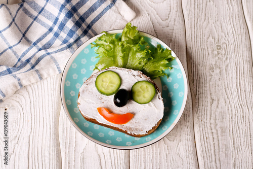 Kids meal, breakfast or lunch for children. Colorful funny food face with healthy vegetables, cheese spread and green salad on a plate. Concept of healthy food, healthy breakfast. Top view, closeup
