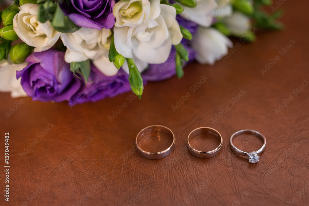 Two golden wedding rings and engagement ring with a diamond on brown background with white and purple flowers Two golden wedding rings and engagement ring with a diamond on brown background with white and purple flowers