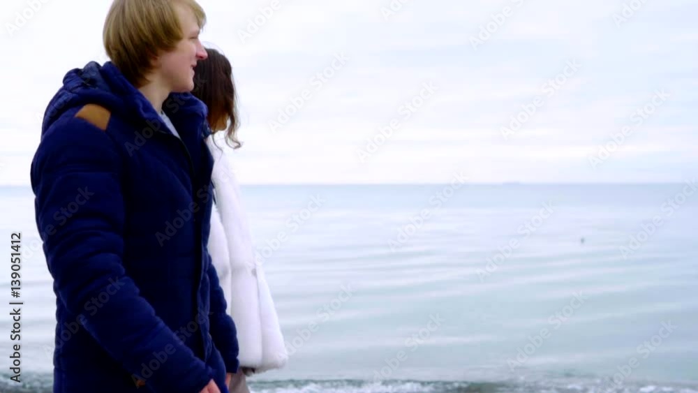 Young couple walking on the beach holding hands. Romantic couple walking wearing coats on sand beach. Slowmotion Young couple walking on the beach holding hands. Romantic couple walking wearing coats on sand beach. Slowmotion