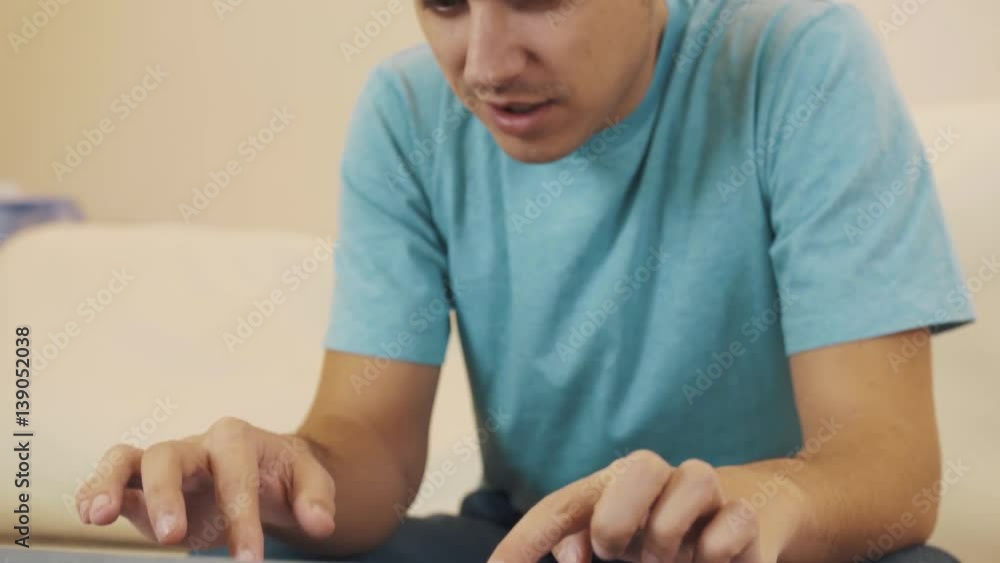 Young caucasian complaining man in blue shirt with hair knot sitting on white leather sofa playing videogame on cased tablet upon glass table Young caucasian complaining man in blue shirt with hair knot sitting on white leather sofa playing videogame on cased tablet upon glass table