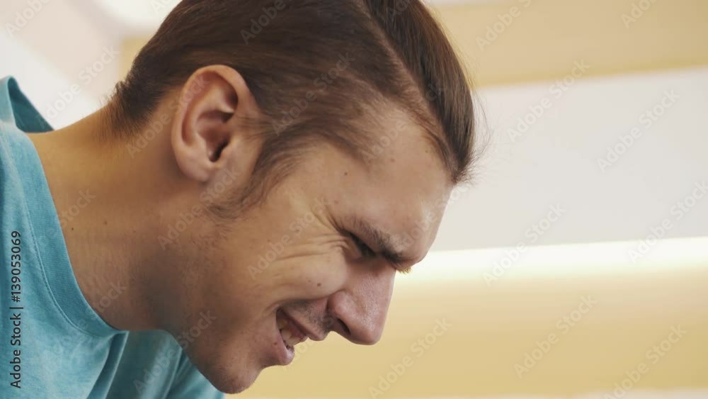Young caucasian smiling man in blue shirt with hair knot sitting on white leather sofa playing videogame on cased tablet upon glass table, speaking and joking. Close up Young caucasian smiling man in blue shirt with hair knot sitting on white leather sofa playing videogame on cased tablet upon glass table, speaking and joking. Close up
