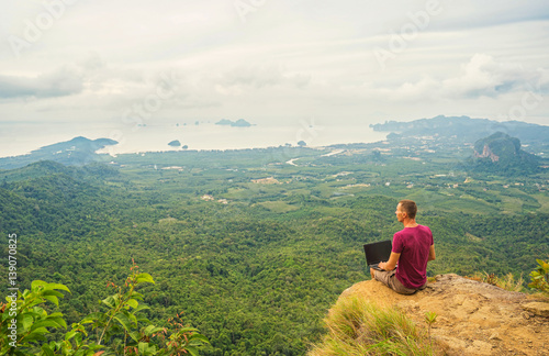 Man working with laptop sitting on the rocky mountain on beautiful scenic clif background. Freelance concept.