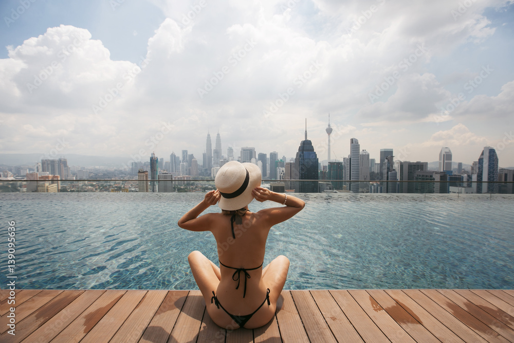Naklejka premium Woman in white hat sitting at Swimming pool on roof top with beautiful city view kuala lumpur Malaysia