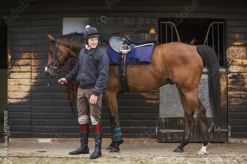 Man wearing riding gear standing outside a box stall at stable with bay horse photo