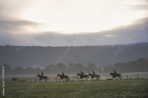 Group of five people on horses riding in a field under a sky with sun and storm clouds. Race horses on the gallops.  photo