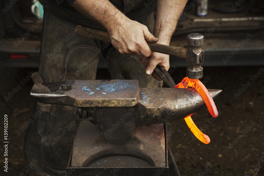 Farrier using tongs and hammer to hold and shape red glowing heated metal horseshoe to be fitted Farrier using tongs and hammer to hold and shape red glowing heated metal horseshoe to be fitted