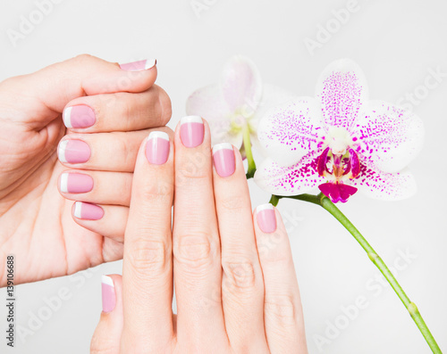 Closeup of female hands and fingers manicured. Fingernails with french spring manicure close to branch of orchid flowers isolated on white background. Painted with modern gel-polish with top cover.