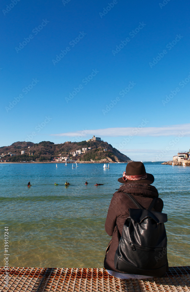 Donostia-San Sebastian, Paesi Baschi, Spagna, 28/01/2017: una ragazza di spalle seduta sul molo con vista sulla spiaggia di La Concha, vista panoramica