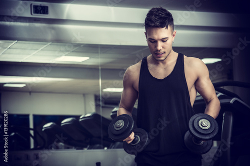 Handsome muscular young man exercising biceps in gym with dumbbells