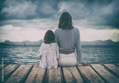 Woman and little girl sitting on the wooden pier near the sea.