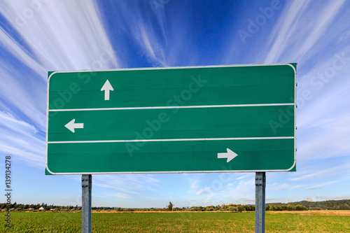 Green road sign with three (3) white arrow in front of a blue sky and green field.