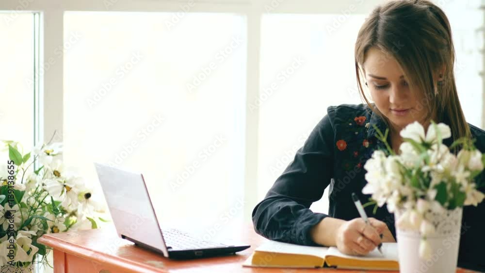 Young beautiful woman student sitting with laptop and writing book at cafe indoors Young beautiful woman student sitting with laptop and writing book at cafe indoors