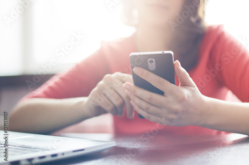 Film effect.  A shot of young female dressed in red casual sweater using high-speed Internet connection on cell phone sitting at the table near her laptop. People, technology and lifestyle concept. photo