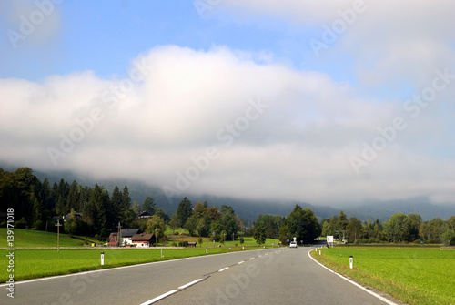 Travel to Sankt-Wolfgang, Austria. The road between green meadows with the mountains in the clouds on the background in the sunny day.