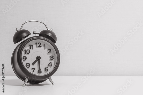 Closeup alarm clock for decorate show half past seven or 7:30 a.m.on white wood desk and cream wallpaper textured background in black and white tone with copy space