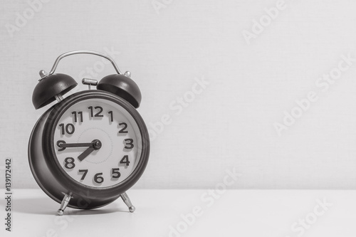 Closeup alarm clock for decorate show a quarter to eight p.m. or 7:45 a.m. on white wood desk and cream wallpaper textured background in black and white tone with copy space