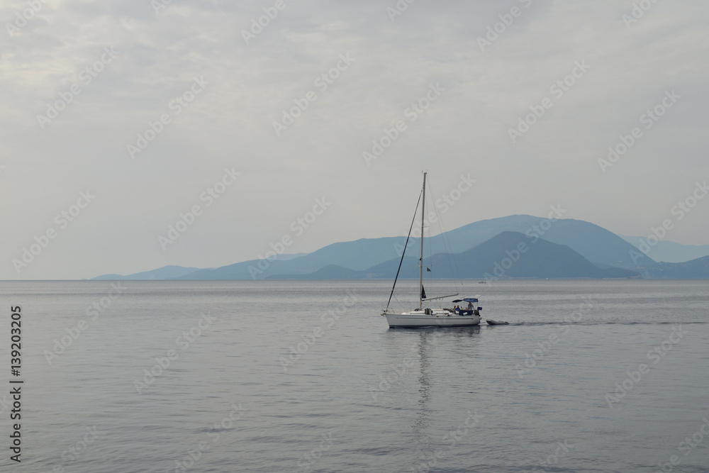 Grey misty day, tranquil sea with smooth surface, yacht in the calm water, silhouette of an island on the horizon