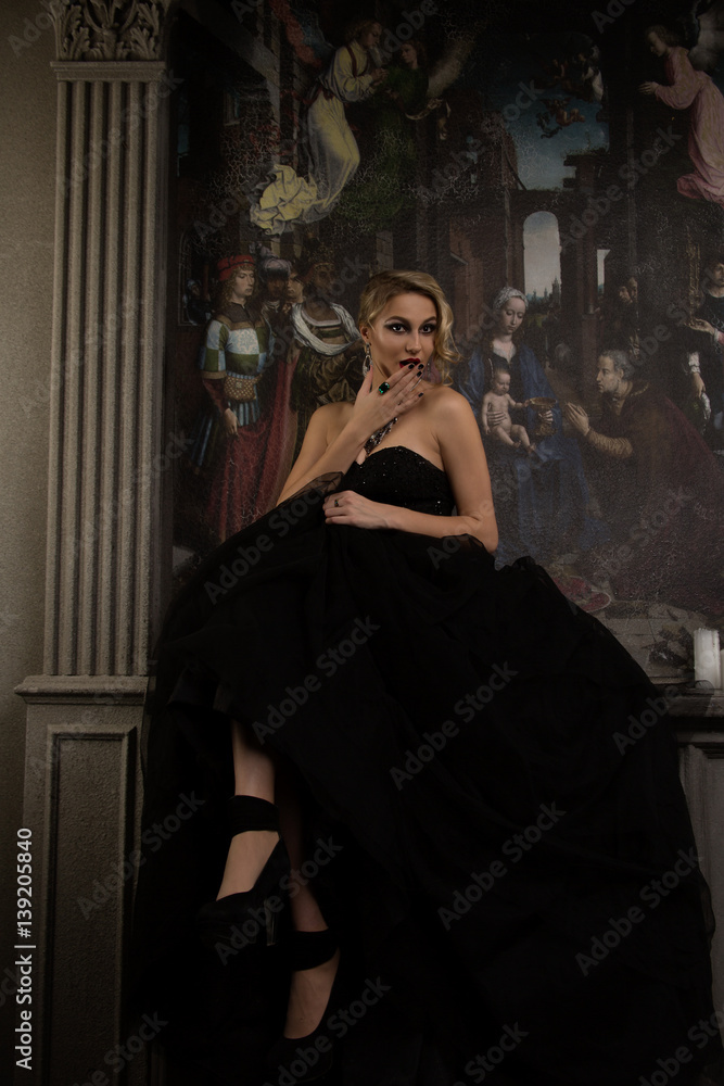 young blond woman sitting on a table in a black ball dress on a background of an arch with a picture Terminated hand over mouth and surprised looks away, at her wearing jewels on her feet black shoes