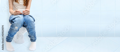 Young woman sitting on bathroom or wc toilet bowl using phone in hands .