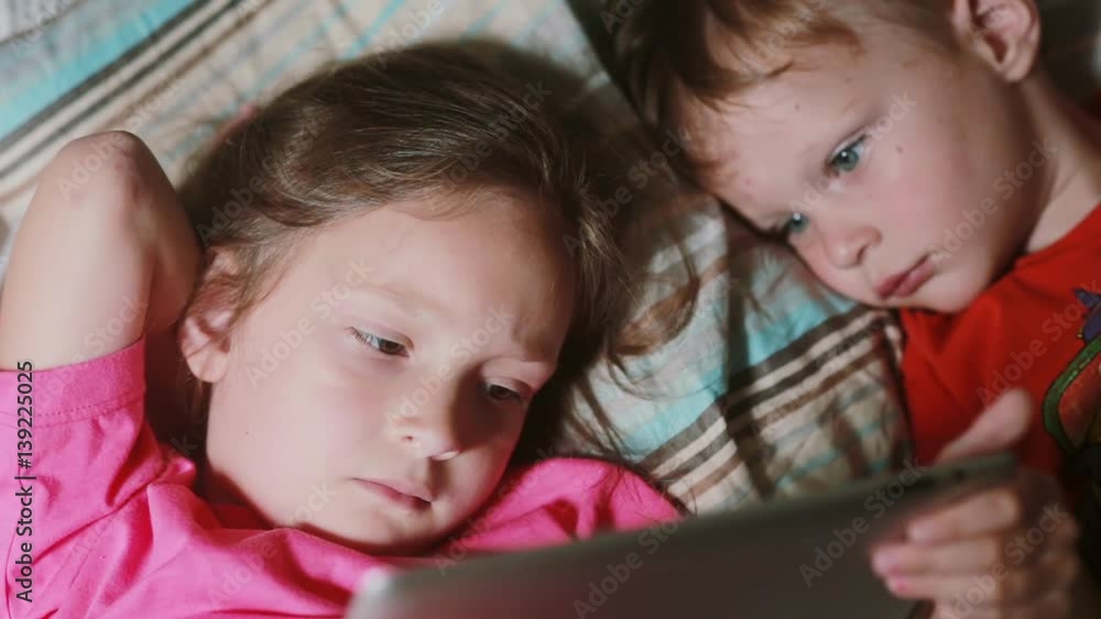 Girl and boy lying on the bed and watching cartoon on touchscreen tablet. Brother and sister having rest together. Girl and boy lying on the bed and watching cartoon on touchscreen tablet. Brother and sister having rest together.