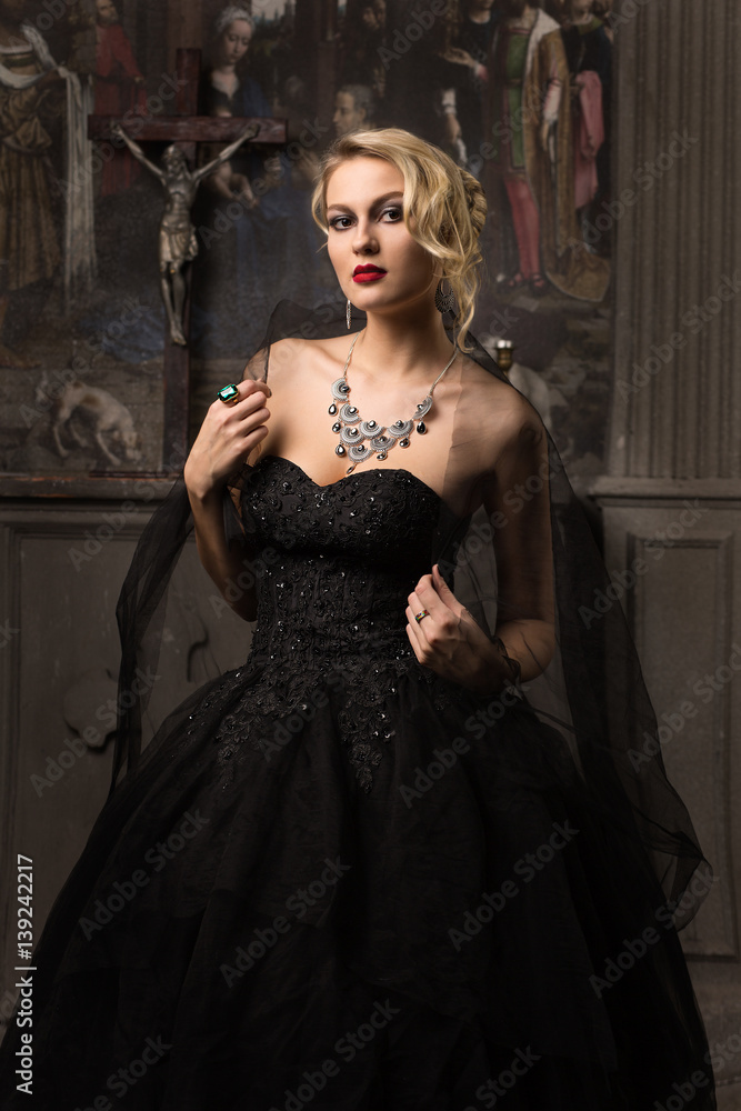 blonde woman in a black evening dress with make-up and hairstyle wearing large jewels veil on his shoulders in the church is the altar on the background and pictures of close-up looks at the camera blonde woman in a black evening dress with make-up and hairstyle wearing large jewels veil on his shoulders in the church is the altar on the background and pictures of close-up looks at the camera