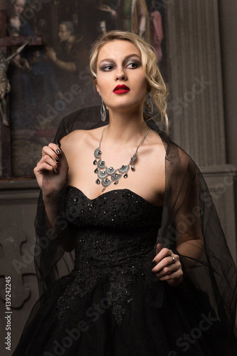 blonde woman in a black evening dress with make-up and hairstyle wearing large jewels veil on his shoulders in the church is the altar on the background close-up looking away from the camera