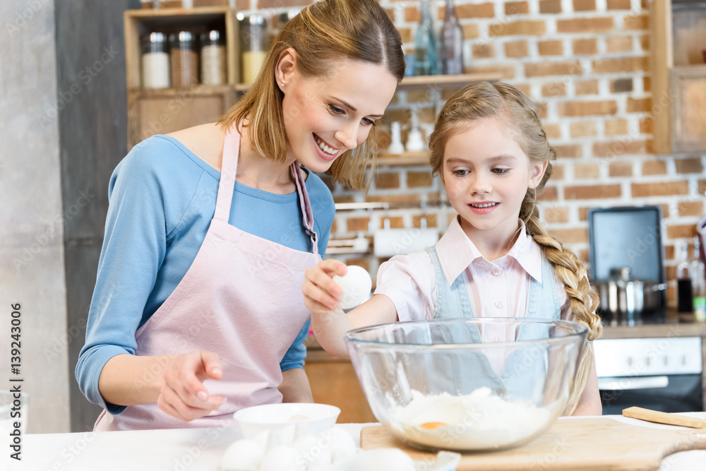 Happy mother looking at daughter beating egg in glass bowl with flour