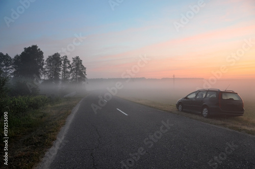 Asphalt road with a parked car in the morning mist on the background of dawn. © struvictory
