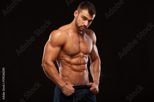 Strong Athletic Man - Fitness Model showing Torso with six pack abs. stands straight and puts his hands in trousers. isolated on black background with copyspace. © satyrenko