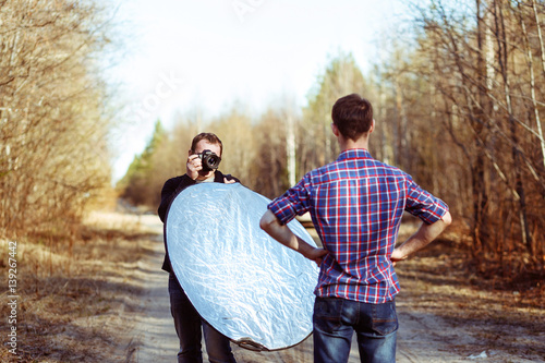 Photographer Photographing Male Model in Forest. Backstage of Fashion Photoshoot by Professional Photographer with DSLR Camera and Reflector photo