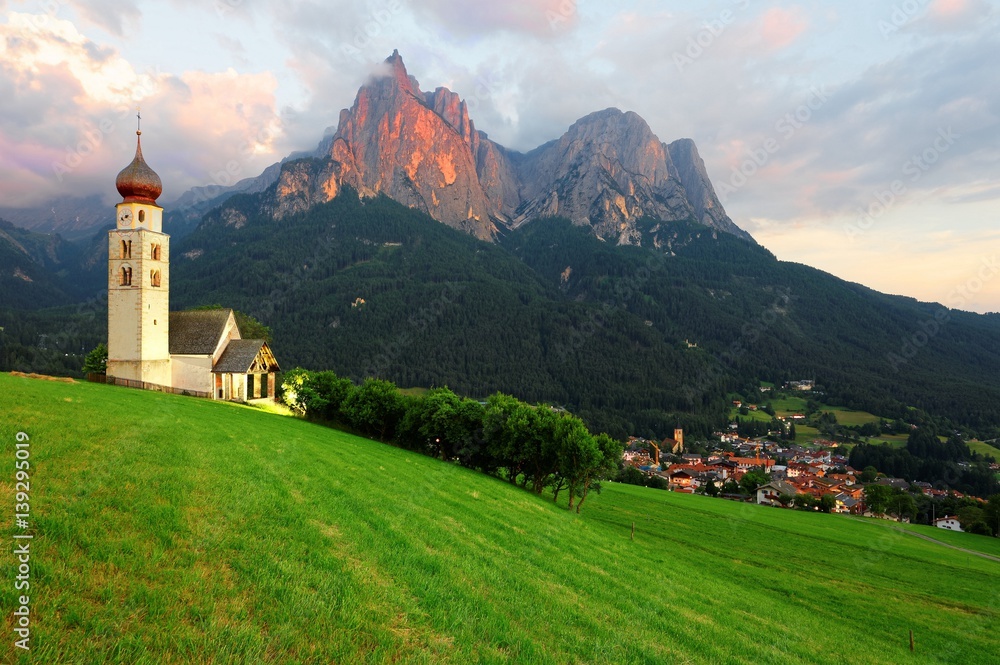 Sunset scenery of Church St. Valentin on green grassy hillside, rugged peaks of Mountain Schlern with alpenglow in background & Village Seis am Schlern in the valley in South Tyrol, Italy, Europe
