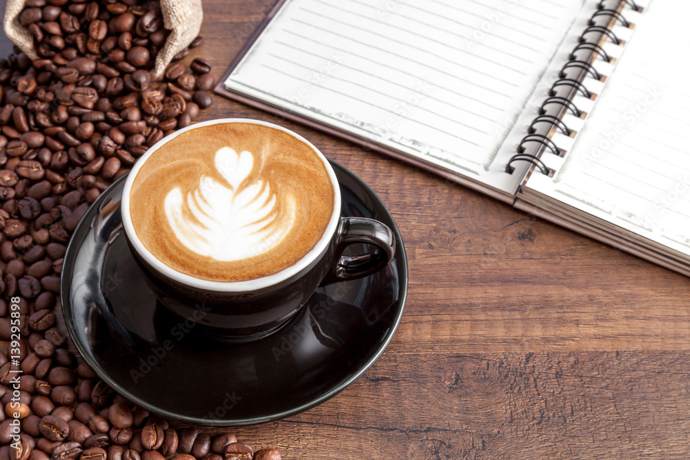 Coffee cup of latte art in the black color cup with some coffee beans and notebook on wooden background with copy space