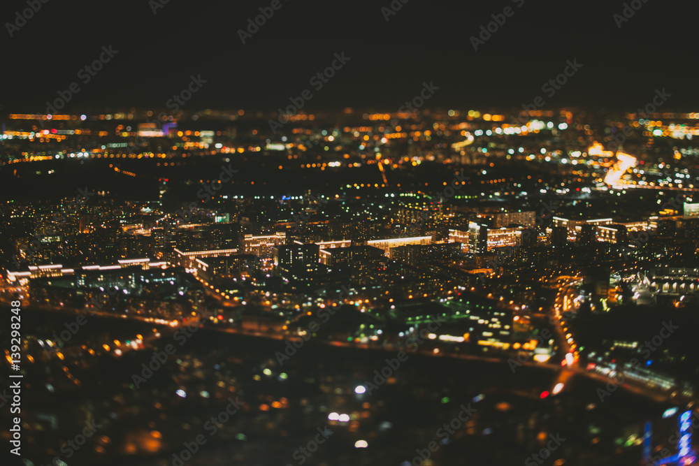 True tilt shift shooting of street surrounded by residential houses of night metropolis from very high point: warm lights from windows, strong bokeh in background and foreground