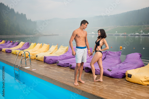 Young romantic couple with perfect bodies standing near swimming pool on the background of beautiful views of forests, hills, river with a haze over it