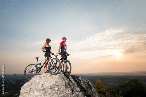 Athletes with mountain bikes standing on the rock under the evening sky and looking into the distance at the sunset