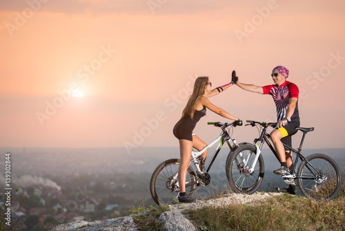 Fototapeta Naklejka Na Ścianę i Meble -  Mountain bikers on the top of a hill, giving a high five to each other against the backdrop of a beautiful sunset. Cyclist standing with the mountain bicycles on the rock