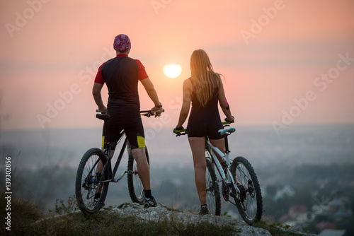 Rear view of guy and girl cyclists on the sports bikes enjoying the beautiful sunset. Bright sun in between. Blurred background.