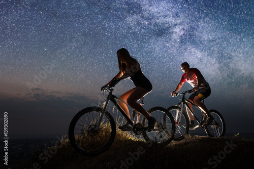 Guy and girl riding a bicycles on the hill at night. Starry sky over the two mountain bikers. Bottom view