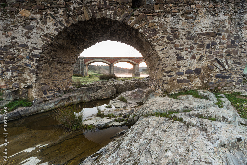 Ancient Roman bridge over the Salor river, located near Aliseda. Extremadura. Spain. Across one of the eyes of the Roman bridge, a modern bridge is seen.