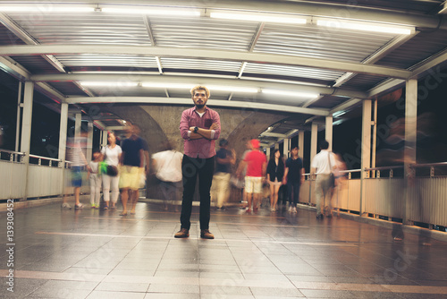 Hipster bearded man standing on the floor with people motion background while traveling at night.