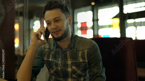 Young man sitting in the dark cafe and chatting on cellphone with someone
