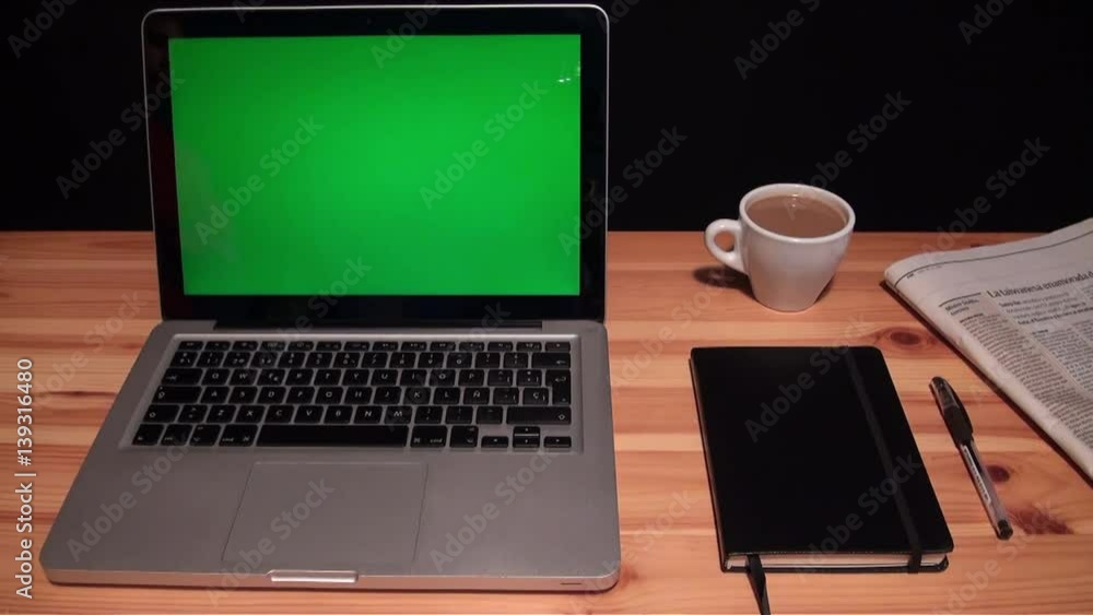 Laptop with green screen chroma on a wooden table and black background. Silver laptop on a wooden table, next door is a coffee, a notebook and a newspaper. Adrian Laptop with green screen chroma on a wooden table and black background. Silver laptop on a wooden table, next door is a coffee, a notebook and a newspaper. Adrian