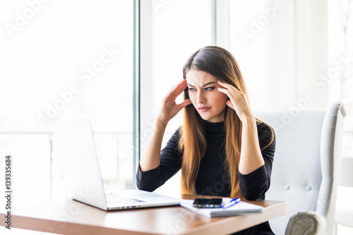 Young attractive woman at modern office desk, working on laptop, massaging temples to forget about constant headaches, noisy loud office giving a migraine, relieving stress, chronic pain, help soothe photo