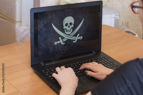 Man or woman hacker or typist, whose face is not visible, holding hands on a laptop keyboard, the screen is a pirate flag Jolly Roger photo