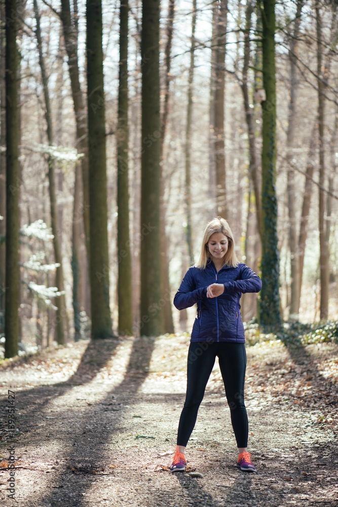 Trail running runner looking at heart rate monitor watch running in forest wearing warm jacket sportswear. Female jogger running training in woods. Trail running runner looking at heart rate monitor watch running in forest wearing warm jacket sportswear. Female jogger running training in woods.