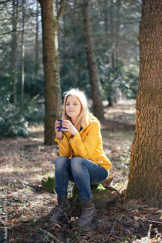 Cute blonde traveler woman resting in forest and drinking hot coffee or tee from a vintage mug